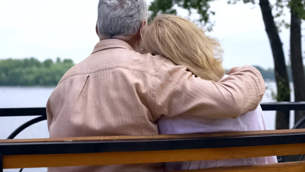 Emerytowany mężczyzna odpoczywa przytulając żonę na ławce w parku w pobliżu rzeki, zdjęcie ilustracyjne